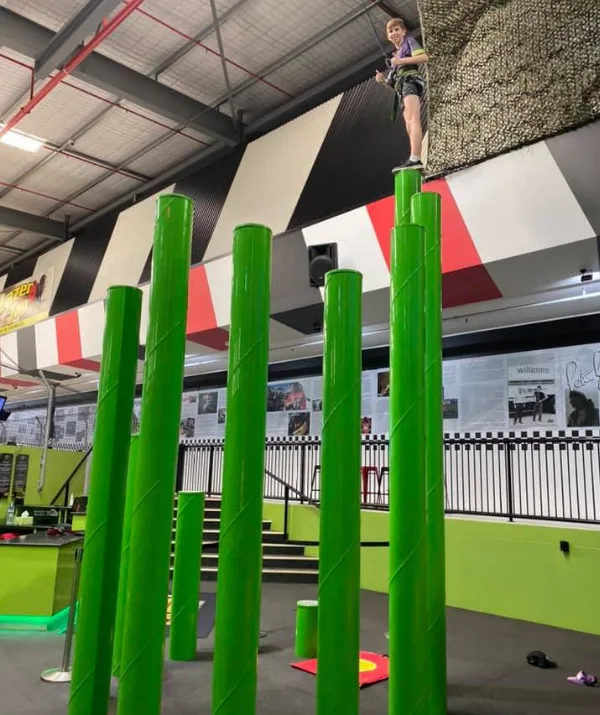Individual stands atop a tall green pole in an indoor activity center, harnessed for safety.
