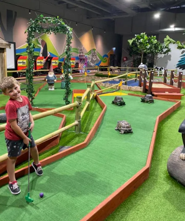 Boy in red shirt smiles while playing mini-golf indoors, with turtles and colorful decorations around the course.