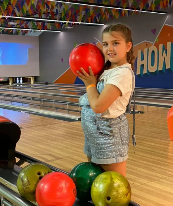 Girl holding a red bowling ball in a colorful bowling alley with pins and balls visible.