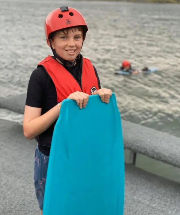 Young person wearing a red helmet and life vest, holding a blue boogie board by a lake.