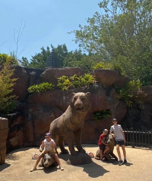 People posing with tiger statues in a landscaped area, surrounded by rocks and greenery.