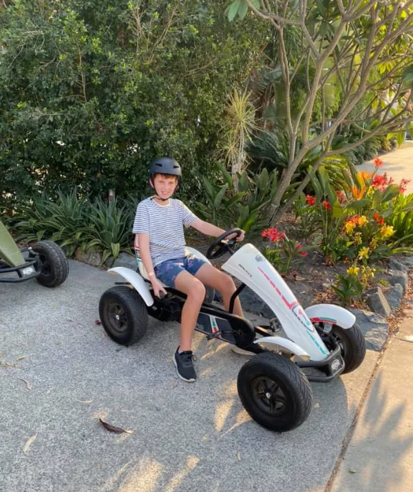 Boy in a helmet sitting on a white go-kart on a paved path with greenery and flowers in the background.