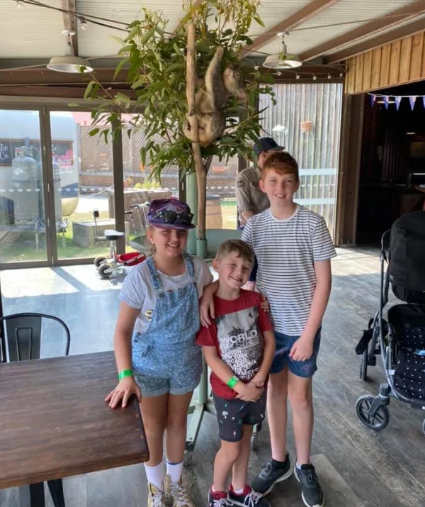 Three children smile indoors near a tree with a sloth hanging from it.