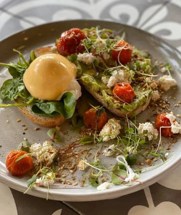 Avocado toast with poached egg, cherry tomatoes, greens, and crumbled cheese on a gray plate.