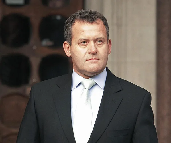 A man in a suit and tie standing outside, looking to the side with a neutral expression.