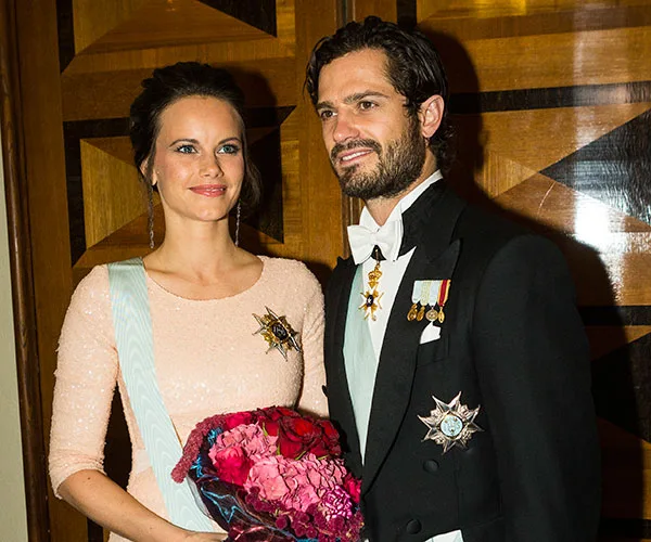 Royal couple in formal attire, the woman holding a bouquet, standing indoors against a geometric wood pattern backdrop.