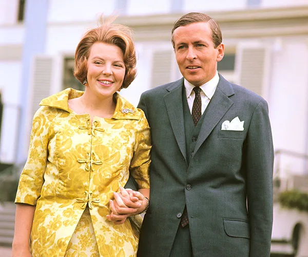 Couple standing, woman in yellow floral dress, man in suit, smiling outdoors.