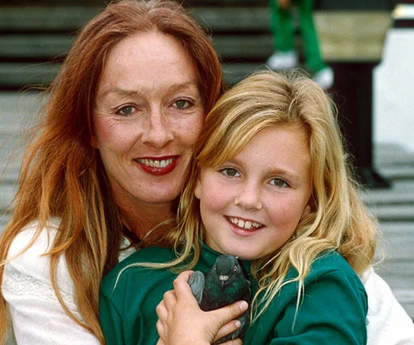 A woman with long red hair hugs a smiling young girl with blonde hair, both posed outdoors.