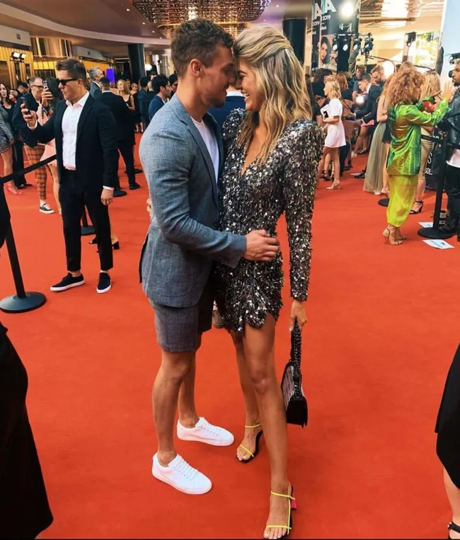 A couple in formal attire shares a moment on a red carpet, surrounded by event attendees and photographers.