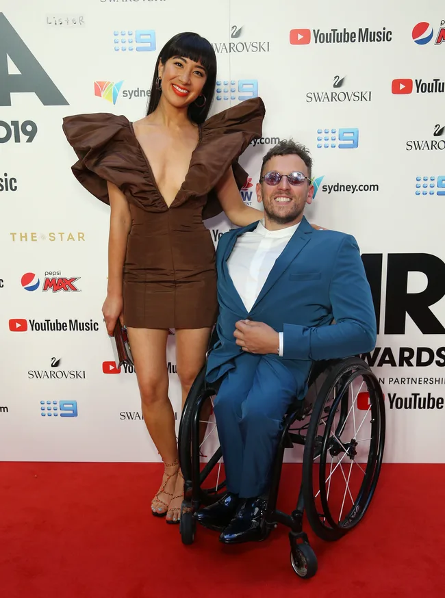 Man in wheelchair wearing blue suit with woman in brown dress on red carpet at an event.