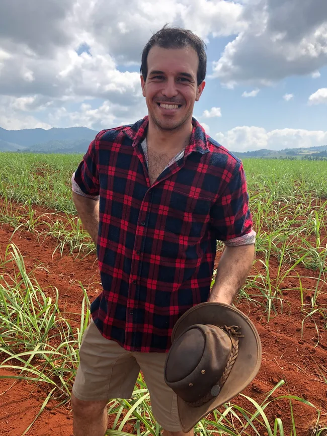 Man in plaid shirt holding a hat, smiling in a field with mountains and a cloudy sky in the background.