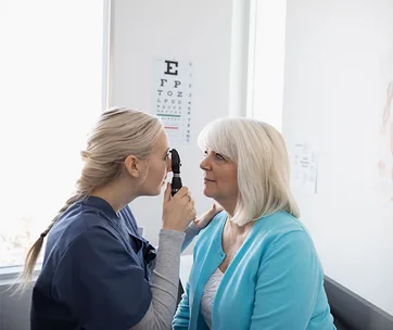 A healthcare professional examines an older woman's eyes in a doctor's office. An eye chart is visible in the background.