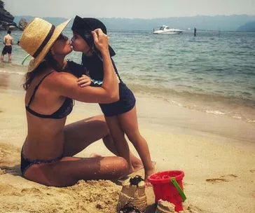 Woman in a straw hat kisses a child on a sandy beach with a red bucket and boat in the background.