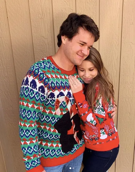 Couple embracing in festive sweaters against a wooden wall, both smiling contentedly.