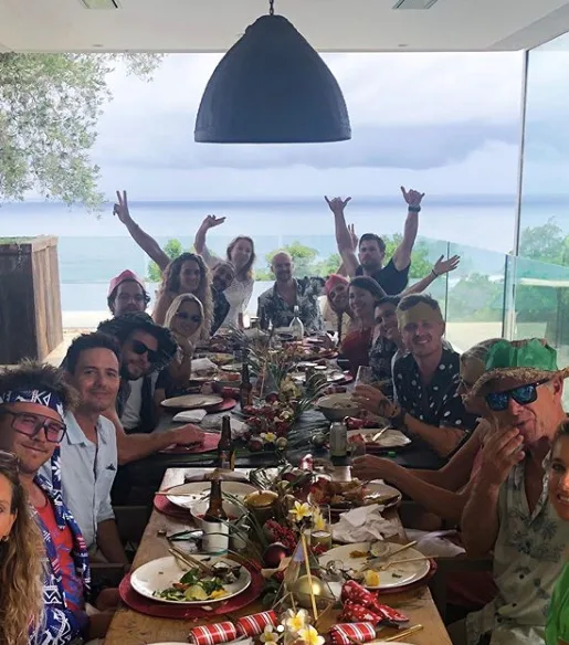 Large group of people sitting around a festive dining table overlooking the sea, smiling and gesturing at the camera.