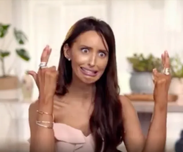 Woman making a funny face with crossed fingers in anticipation, wearing jewelry and a pink outfit indoors.