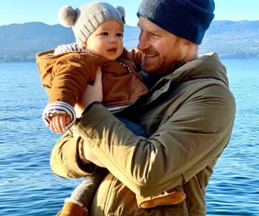 A man in a beanie holds a baby in a brown jacket by a lake with mountains in the background.