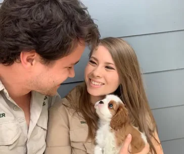 Couple smiling at each other while holding a small brown and white puppy indoors.