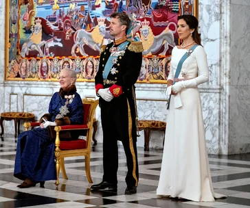 Three royals in formal attire, including Princess Mary in a white dress, stand in a grand hall with a colorful tapestry.