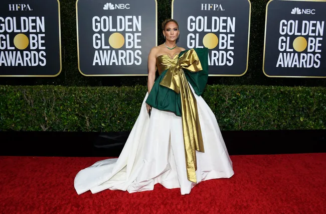 Person in an elegant white gown with a large gold and green bow stands on a red carpet at the Golden Globe Awards.