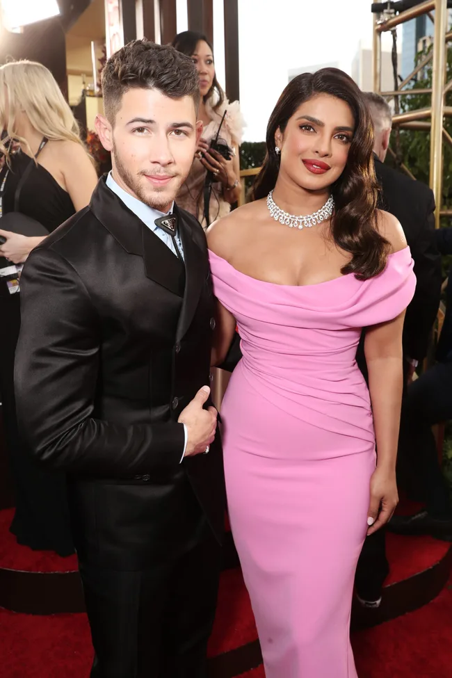 A well-dressed couple poses on the red carpet; the woman wears a pink gown and diamond necklace.