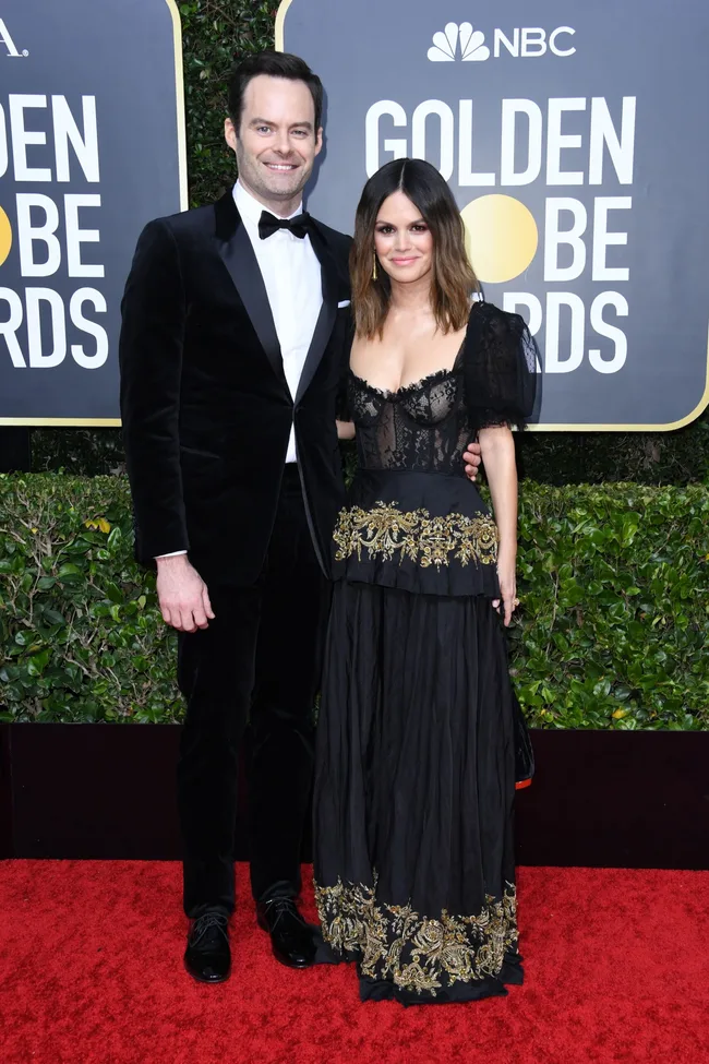 Man in black tuxedo and woman in black gown with gold accents on red carpet at Golden Globe Awards.