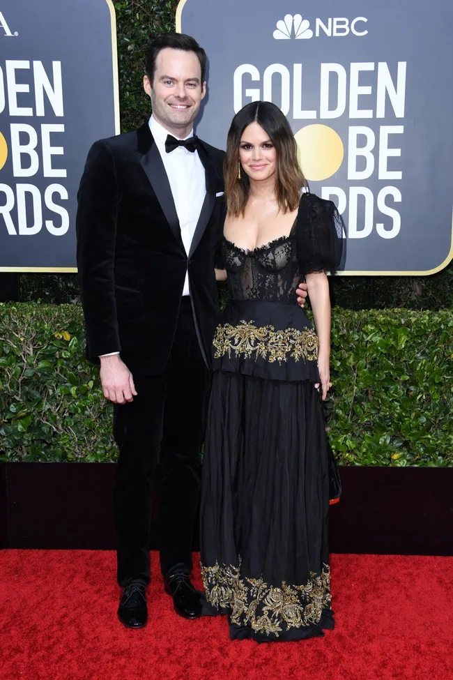 Man in black tuxedo and woman in black gown with gold accents on red carpet at Golden Globe Awards.