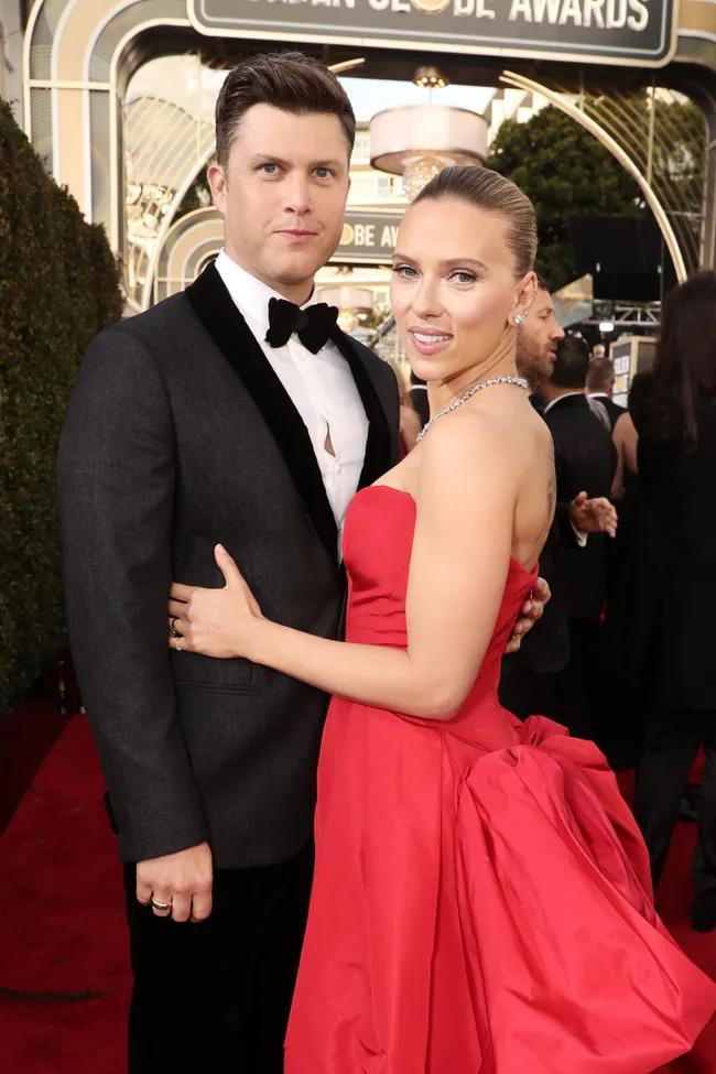 Couple poses on the red carpet at an awards event; a man in a tuxedo and a woman in a red gown.