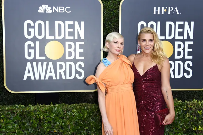 Two individuals posing in front of Golden Globe Awards signage; one in an orange dress, the other in a red sequined dress.