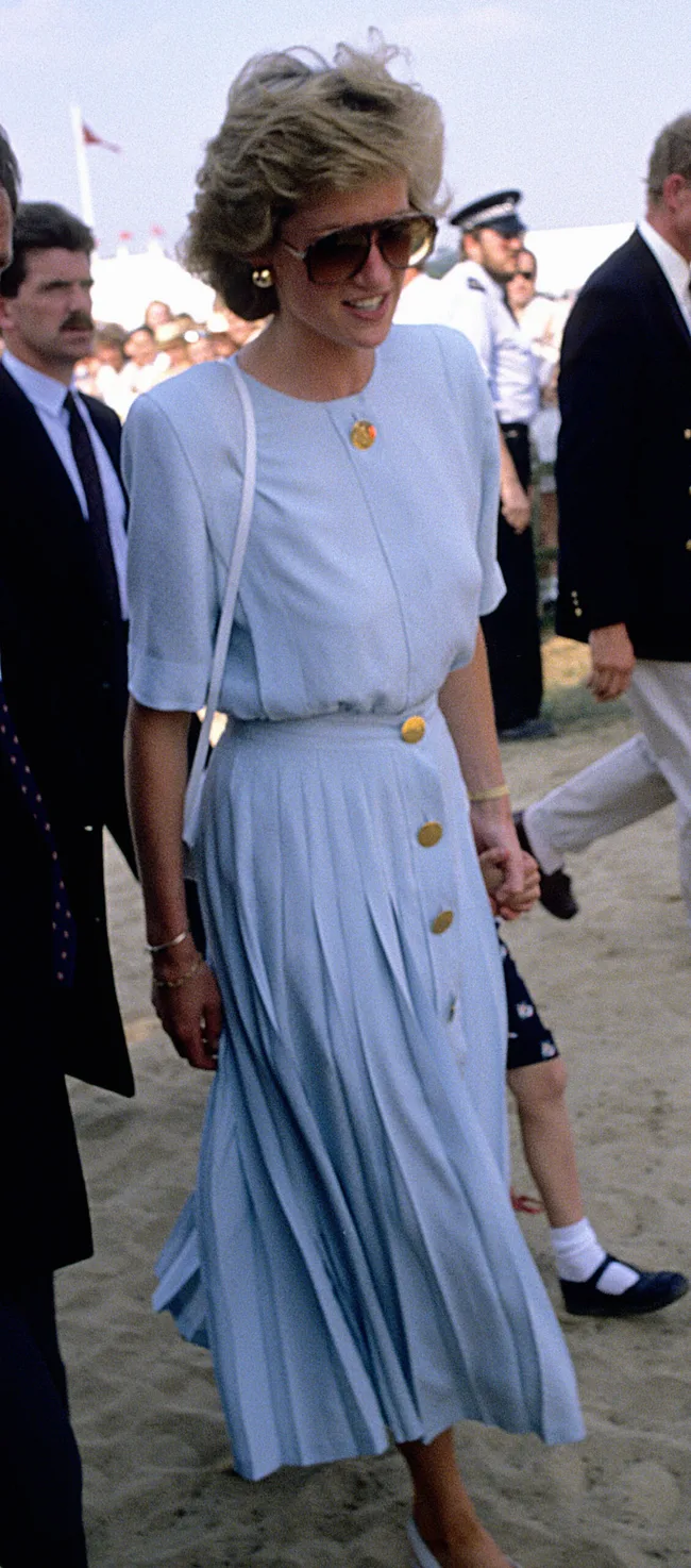 A woman in a light blue dress, sunglasses, and a white purse walks outdoors, surrounded by people.