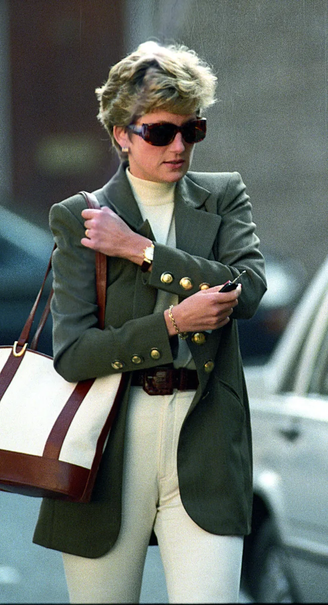 A person in sunglasses and a green blazer carrying a large tote bag, holding a key, walking on a city street.