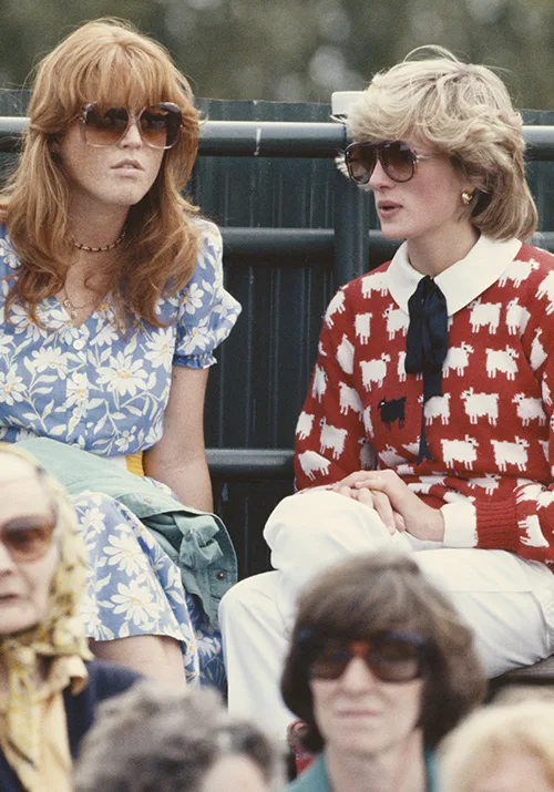Two women sitting outdoors wearing sunglasses; one in a floral dress, the other in a sheep-patterned sweater.