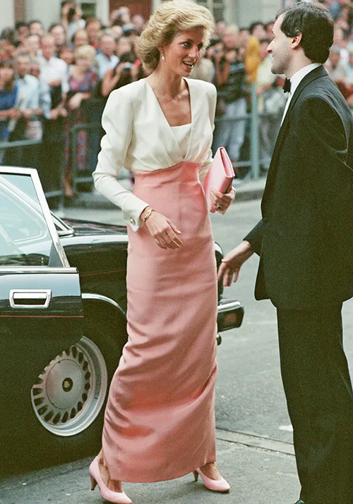 Woman in formal pink and white attire smiling by a car, holding a clutch, with people in the background.