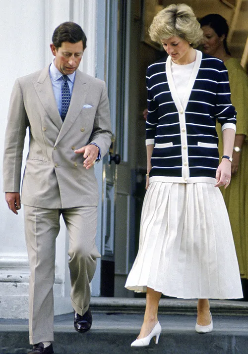 A man in a beige suit and a woman in a striped cardigan and white skirt walk down steps side by side.