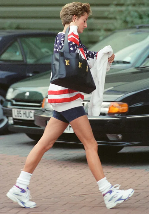 A person walking, wearing an American flag sweater and shorts, carrying a black bag and a white item, with cars in the background.