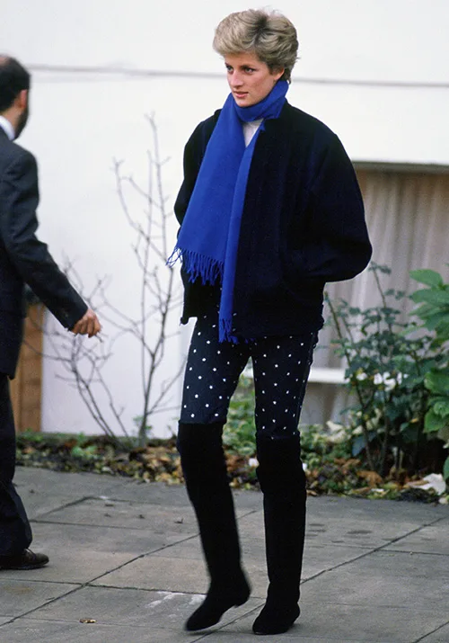 Person wearing a blue scarf, dark jacket, polka dot pants, and black boots, walking on a paved path.