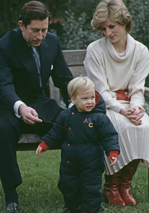 A young boy in a navy coat stands on grass, with two adults seated on a bench behind him in an outdoor setting.