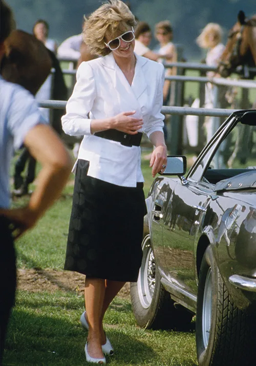 Elegant woman in a white jacket and black skirt, wearing sunglasses, smiling beside a classic car at an outdoor event.