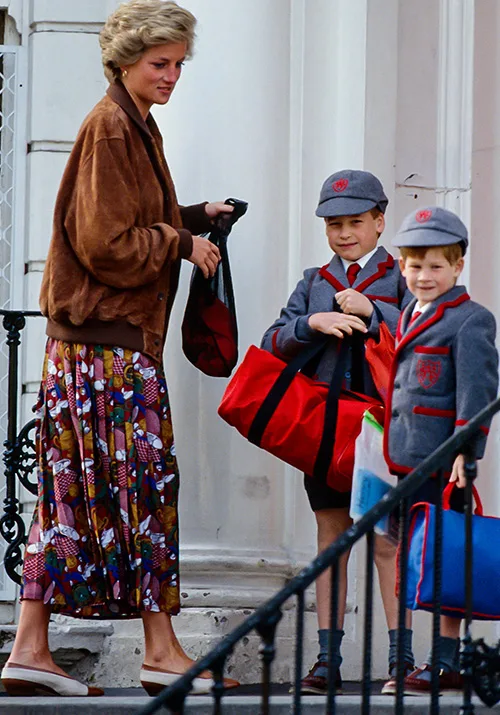 Woman with short hair escorts two boys in school uniforms carrying bags on steps of a building.
