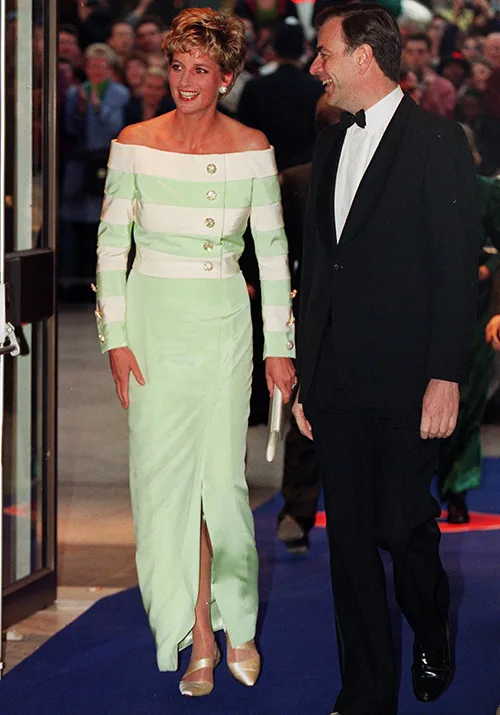 A woman in a green and white off-shoulder dress walks on a blue carpet with a man in a tuxedo, surrounded by a crowd.