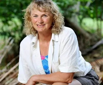 A woman in a white shirt sitting outdoors with a blurred natural green background, smiling at the camera.