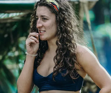 A woman with curly hair and a headband, wearing a blue top, appears thoughtful in an outdoor setting.
