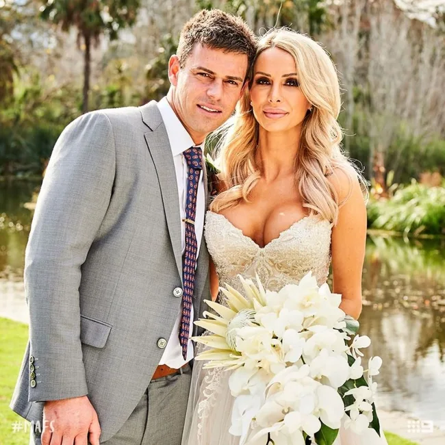 A bride and groom pose outdoors by a pond, with the bride holding a bouquet of white flowers.
