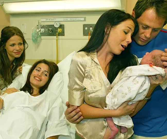 A hospital scene with three women and one man celebrating a newborn baby wrapped in a blanket.