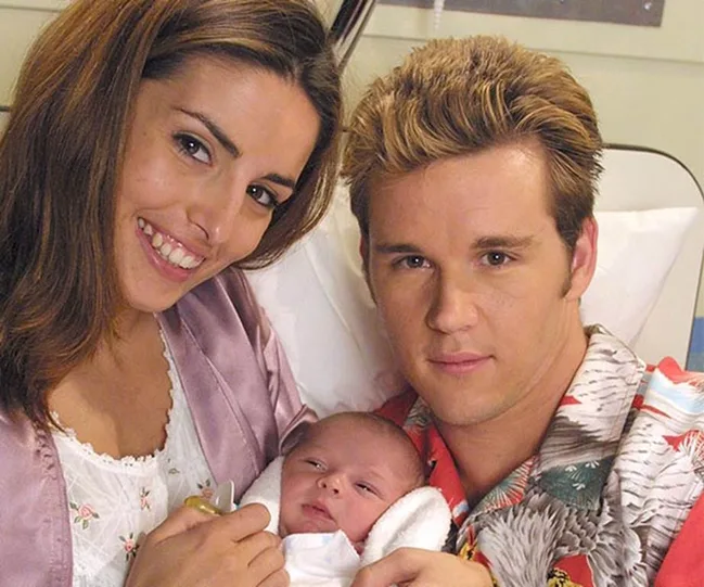 A man and woman smiling while holding a newborn baby, with the woman wearing a light robe and the man in a colorful shirt.