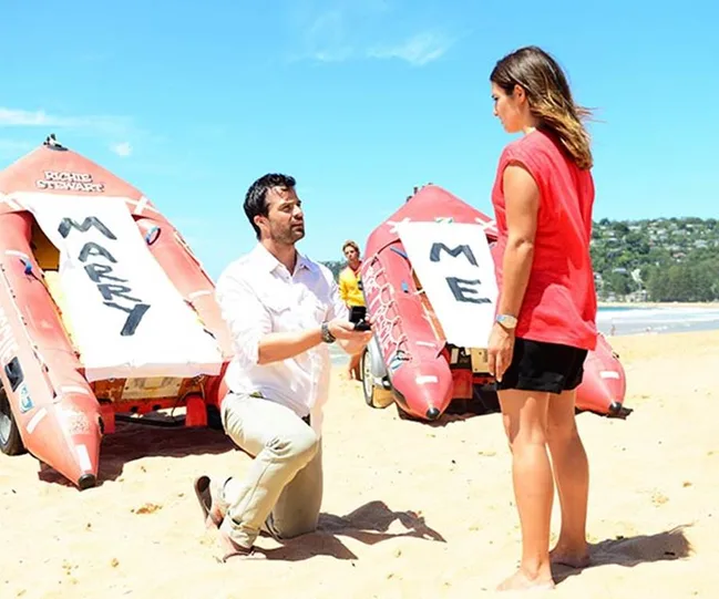 Man proposing on beach with "Marry Me" signs on boats behind him, holding ring towards woman.