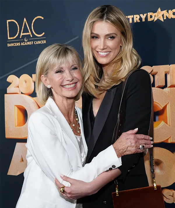 Two smiling women embracing each other at an event, with "Dancers Against Cancer" signage in the background.