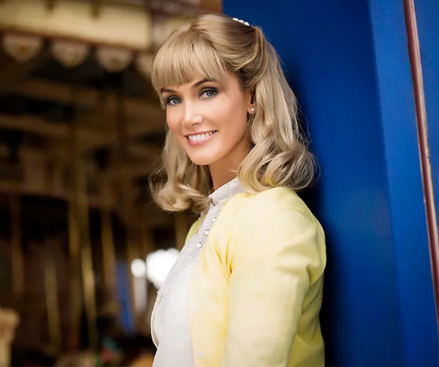 Smiling woman with blonde hair in a yellow cardigan, leaning against a blue wall.