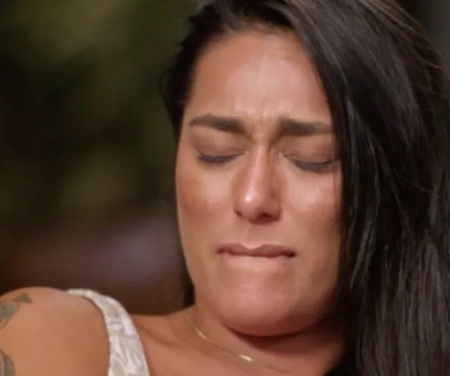 Woman with long dark hair, eyes closed, appears emotional, biting lip, wearing a white top.