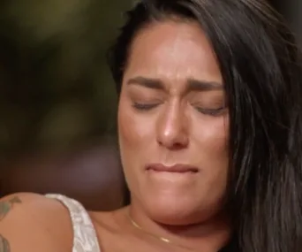 Woman with long dark hair, eyes closed, appears emotional, biting lip, wearing a white top.